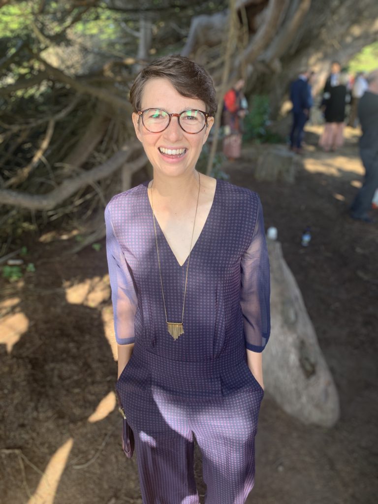 Abby wearing a plum-colored jumpsuit and smiling in the foreground. Nature scene in the background.