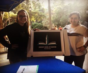 This image shows two women standing on each side of a sign reading "Graduate School of Education Research Day" with an image of an open book and the Campanile behind.  
