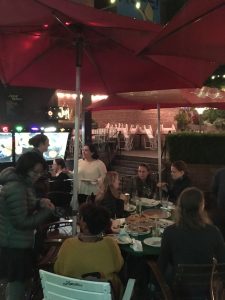A group of people sit around a table with pizza and plates beneath red umbrellas